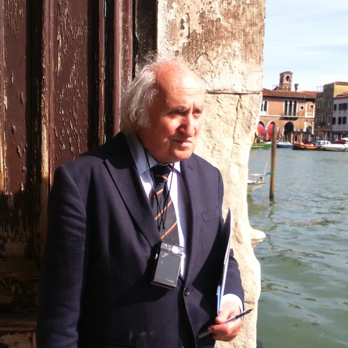 Richard Demarco in a suit stands by a weathered stone wall near a Venetian canal, holding documents and wearing a device around his neck. Boats and historic buildings are visible in the background.